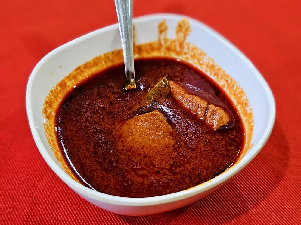 A white bowl of dark red mutton curry on a bright red tablecloth, a spoon resting in the gravy