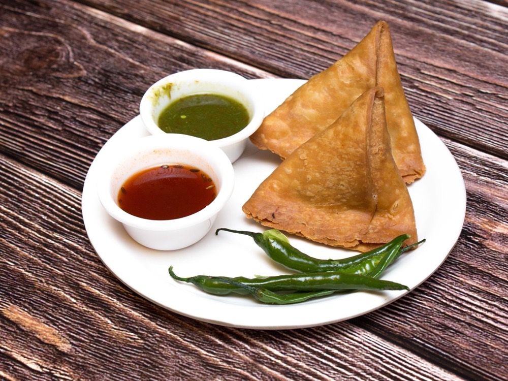 Two golden samoosas on a white plate with green chutney and red sauce in small bowls, and two green chillies